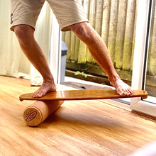 Load image into Gallery viewer, A person standing on a balance board with a cork roller attached to it, positioned on a wooden floor.